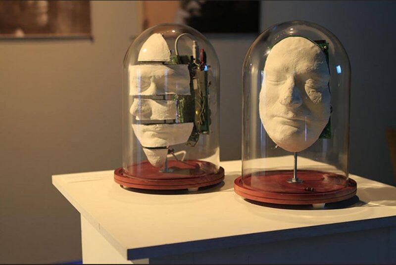 Two sculpted faces attached to motherboards are on display in glass jars
