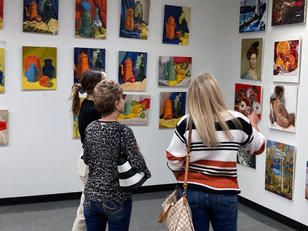 Three people look at a wall of paintings at an art exhibit.