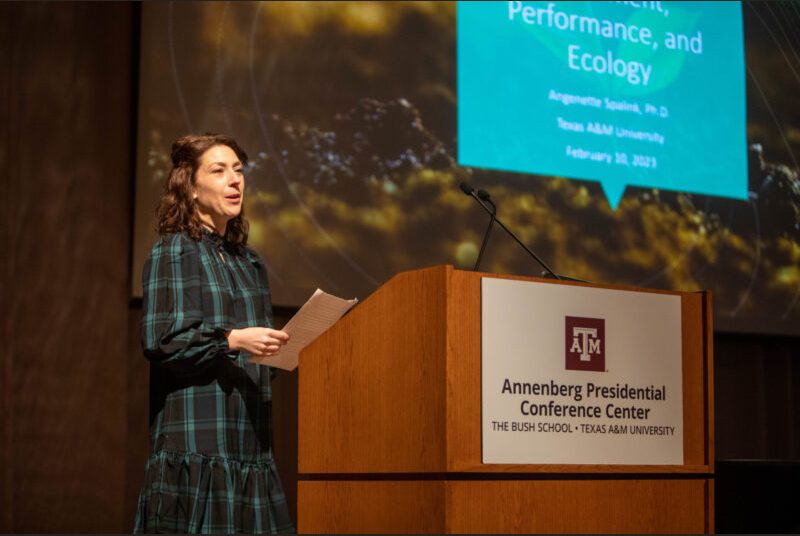 A woman stands at a podium giving an academic presentation