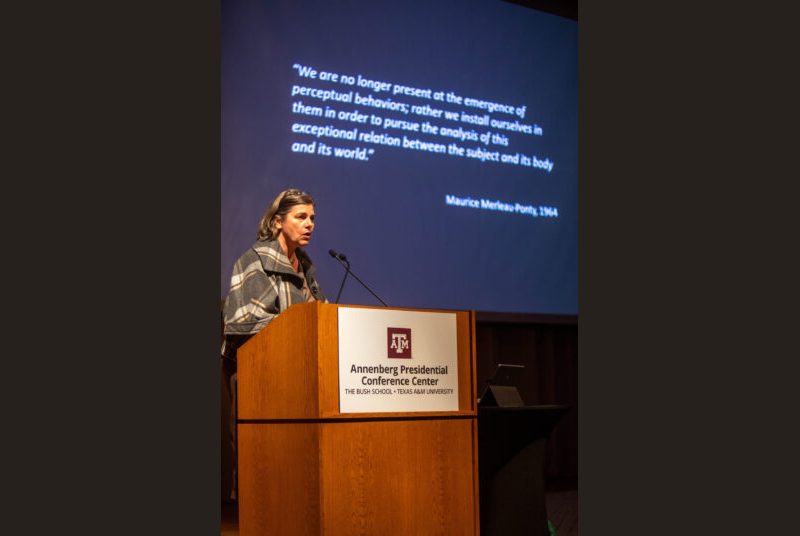 A woman stands at a podium giving an academic presentation