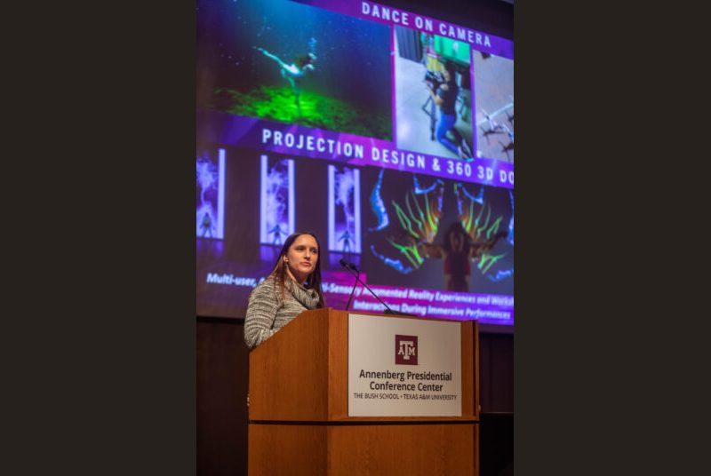 A woman stands at a podium giving an academic presentation