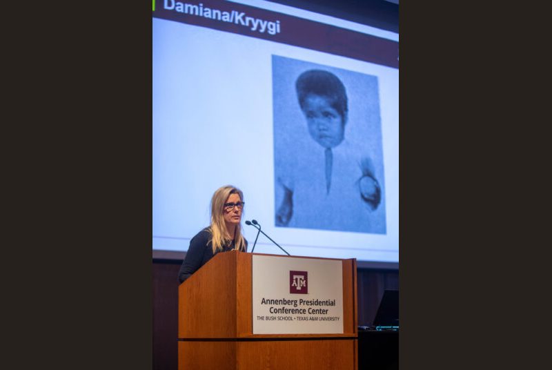 A woman stands at a podium giving an academic presentation