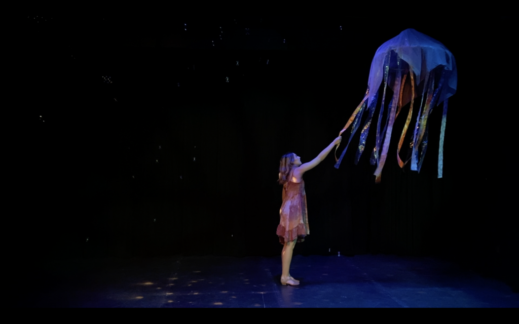 A performer onstage reaches out and grasps a ribbon suspended from the ceiling.