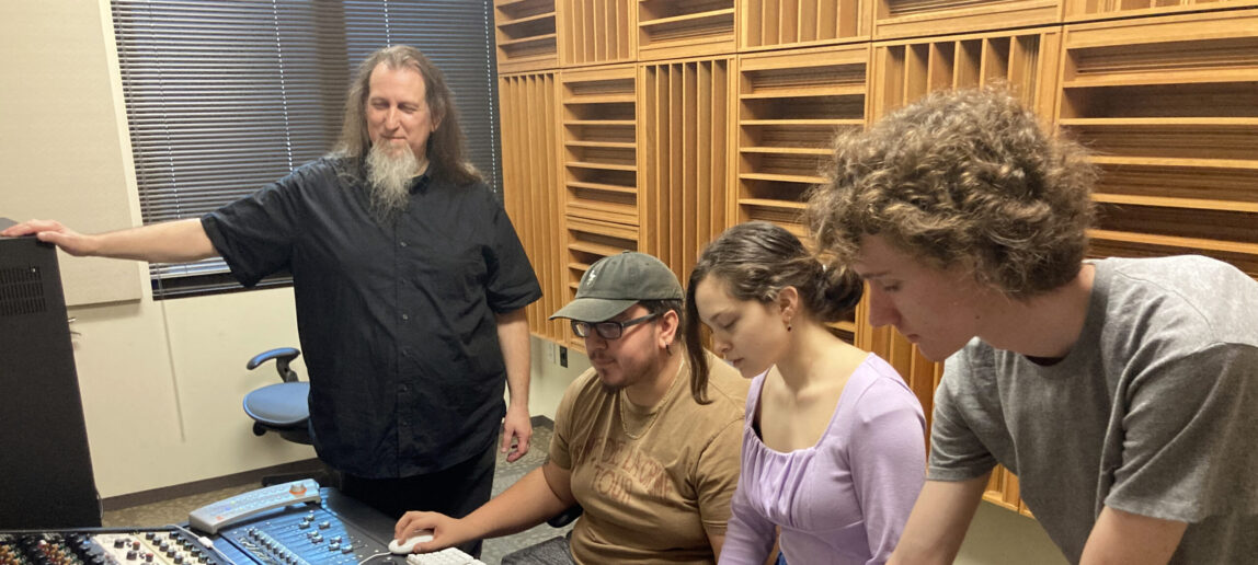 A lecturer stands by three college students who are working with recording studio equipment.