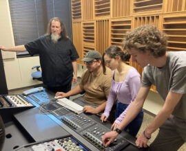 A lecturer stands by three college students who are working with recording studio equipment.