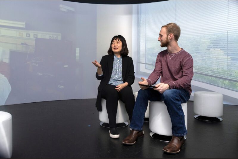 A professor speaks with a student while seated inside a room with a 360-degree screen.