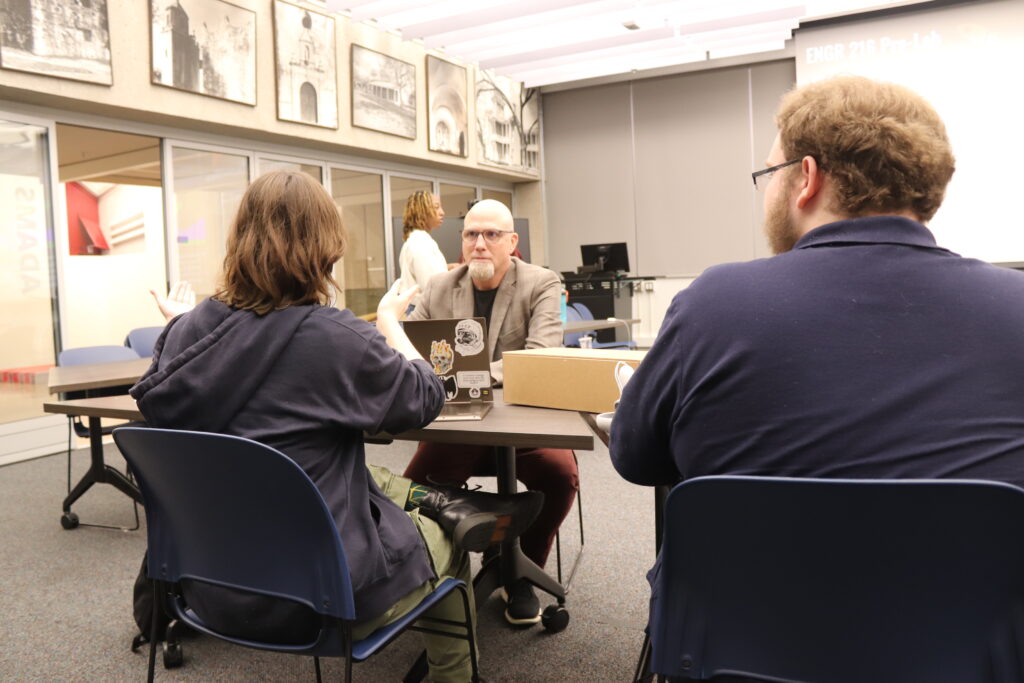 A man facing the camera view sits at a table opposite two college students as they discuss virtual reality projects.