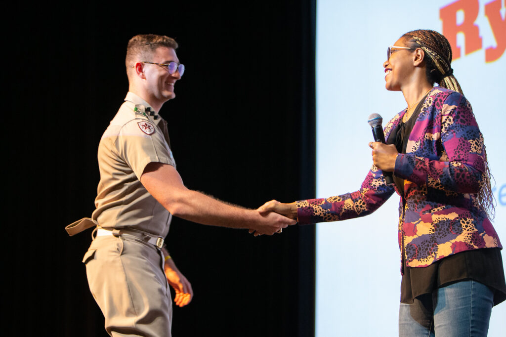 A college student in a khaki ROTC uniform shakes the hand of a professor onstage