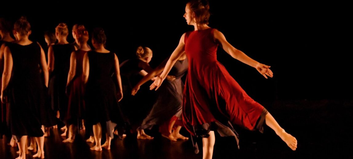 A dancer in a red dress stands on one foot and gestures with her arms as a group of other dancers in black have their backs turned.