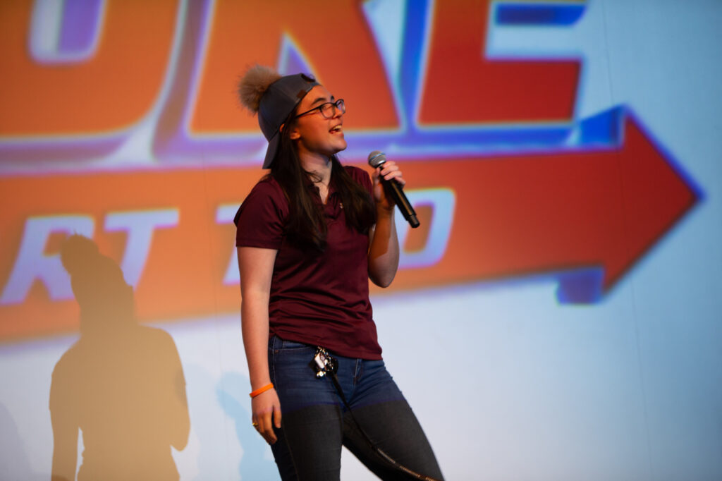 A college student wearing a backward cap stands onstage and speaks with a microphone to an audience.