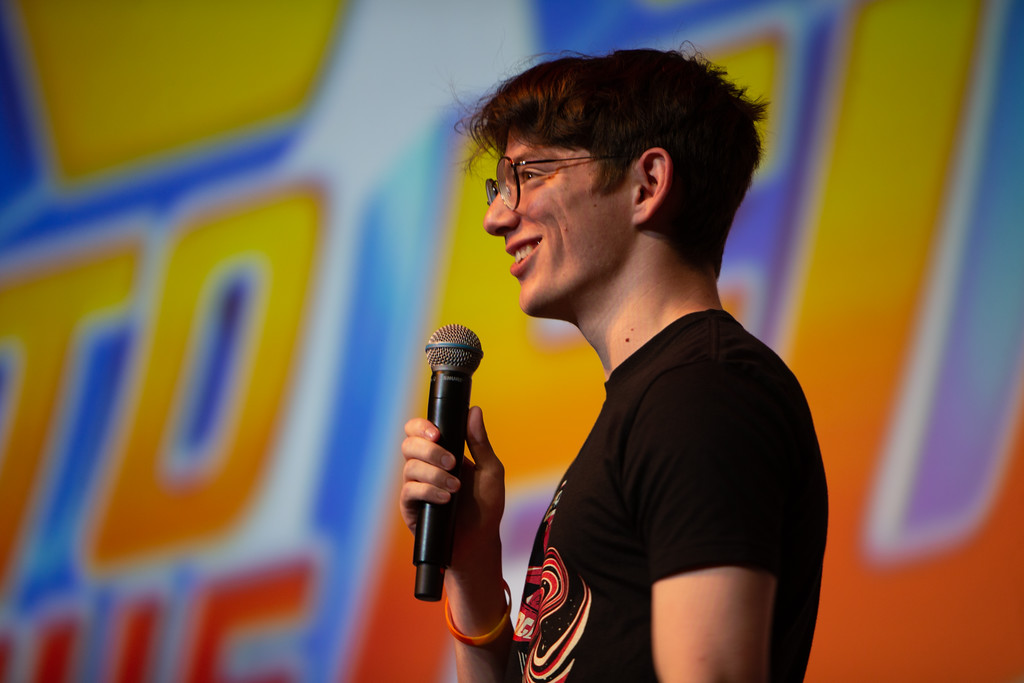 A man wearing glasses and a black T-shirt stands onstage and speaks to an audience using a microphone.