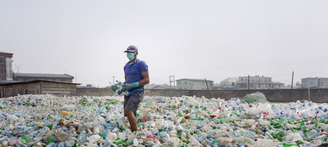 A man with a face mask on walks amid an enormous pile of empty plastic water bottles.