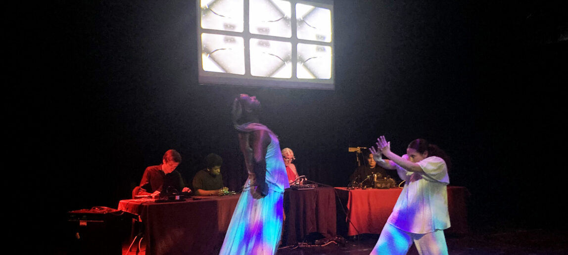 Two dancers perform in front of four people playing electronic instruments.