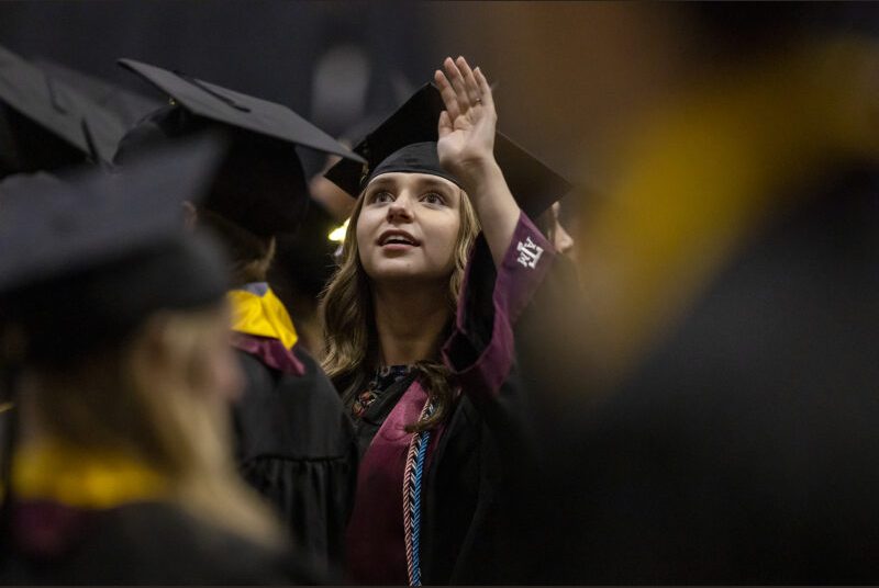 A graduating college student looks up into the audience at a graduation ceremony.
