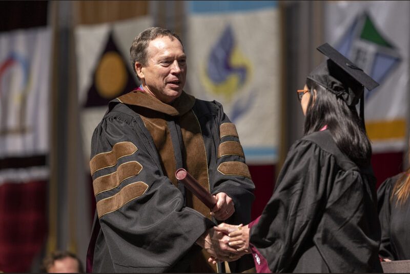 A school leader gives a diploma to a college student wearing a black cap and gown.