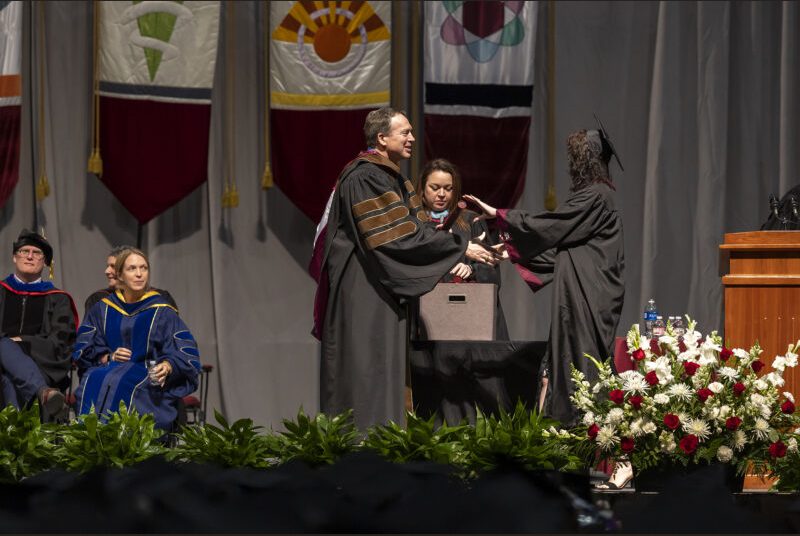 A school interim dean gives a diploma to a college student wearing a black cap and gown.
