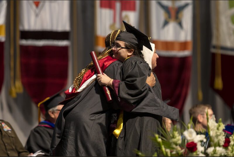 A college professor hugs a new college graduate wearing a black cap and gown.