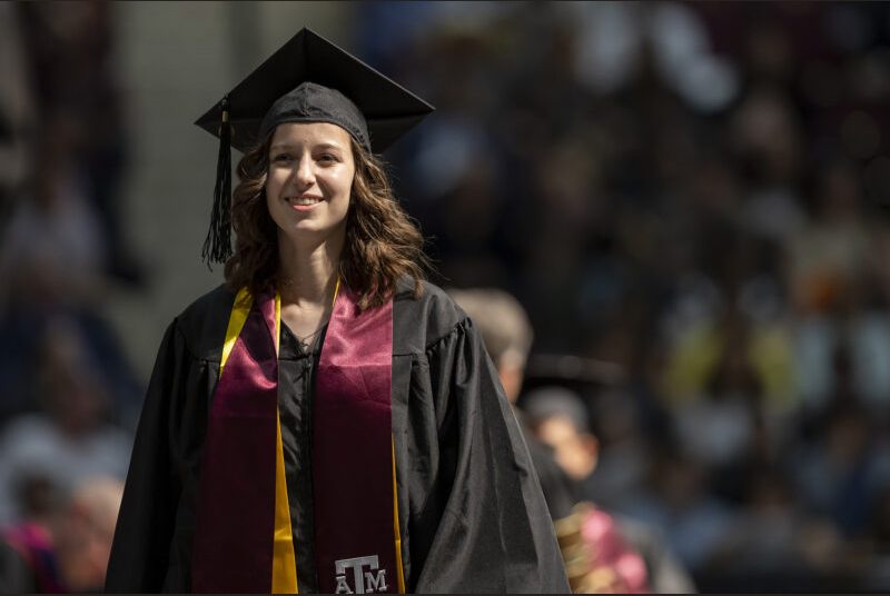 A new college graduate wearing a black cap and gown with maroon and yellow sashes receives her diploma at a graduation ceremony.