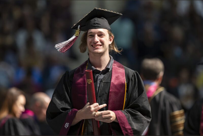 A new college graduate wearing a black cap and gown with a maroon sash walks across the stage at a graduation ceremony.