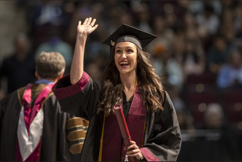 A new college graduate wearing a black cap and gown with maroon and yellow sashes walks across the stage at a graduation ceremony.