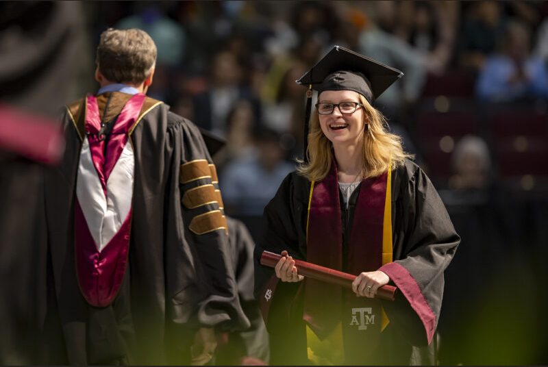 A new college graduate wearing a black cap and gown with maroon and yellow sashes received her diploma at a graduation ceremony.