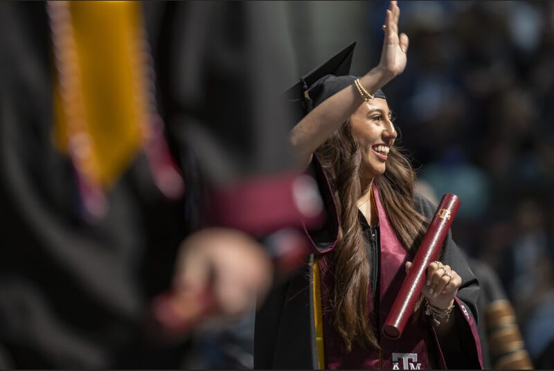 A new college graduate wearing a black cap and gown with a maroon sash waves after receiving her diploma at a graduation ceremony.