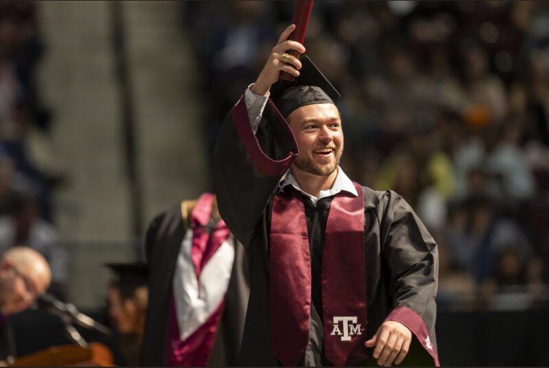 A new college graduate wearing a black cap and gown with a maroon sash receives his diploma at a graduation ceremony.