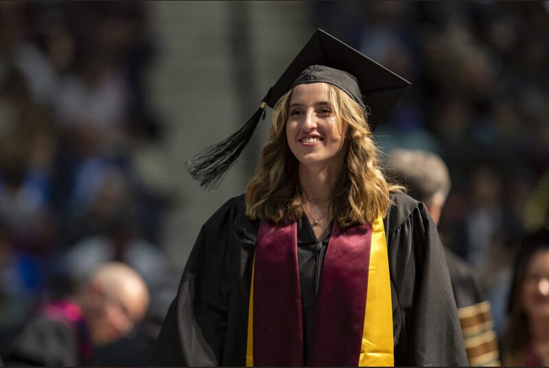 A new college graduate wearing a black cap and gown with maroon and yellow sashes walks across the stage at a graduation ceremony.