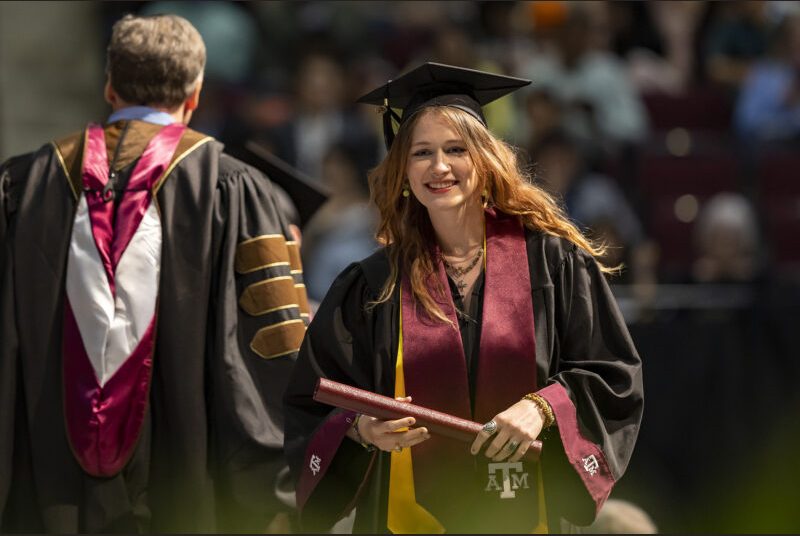 A new college graduate wearing a black cap and gown with maroon and yellow sashes receives her diploma at a graduation ceremony.