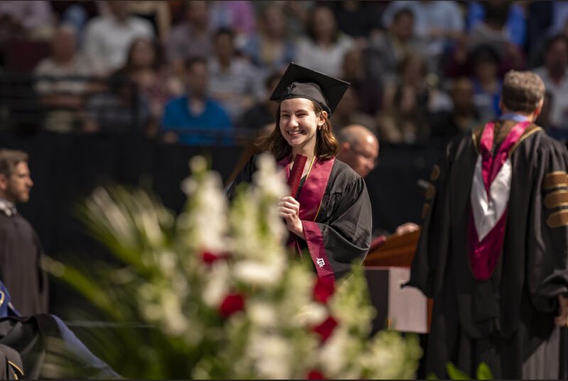 A new college graduate wearing a black cap and gown with a maroon sash receives her diploma at a graduation ceremony.
