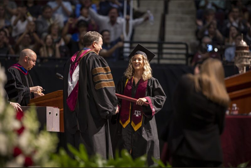 A new college graduate wearing a black cap and gown with maroon and yellow sashes received her diploma at a graduation ceremony.