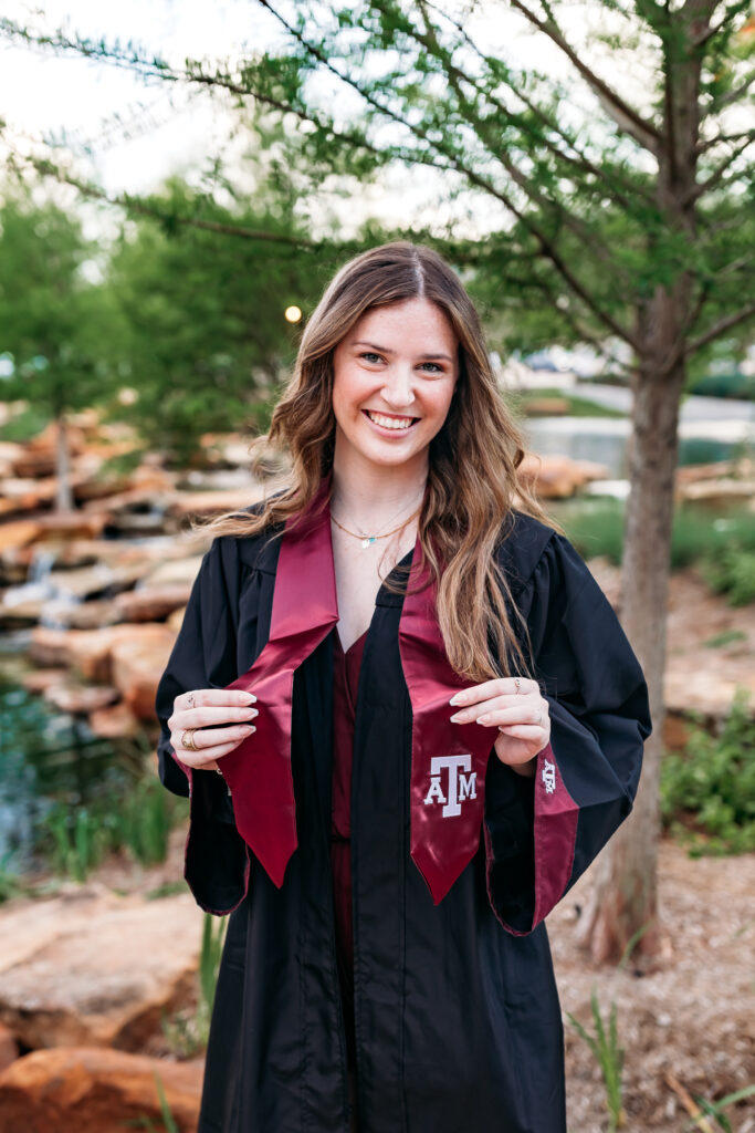 A college student wearing a graduation gown and maroon sash smiles in an outdoor setting.
