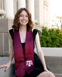 A college student wearing a maroon graduation sash sits outside of a building.