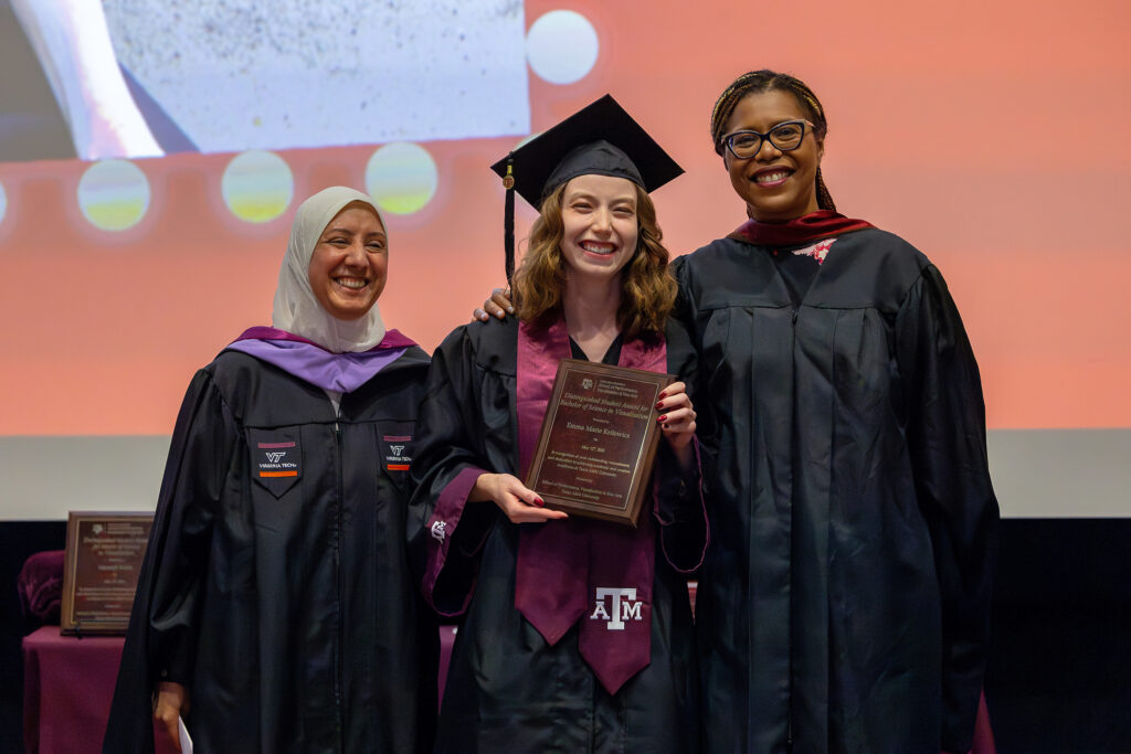 A college student in a graduation cap and gown stands between two college professors onstage.