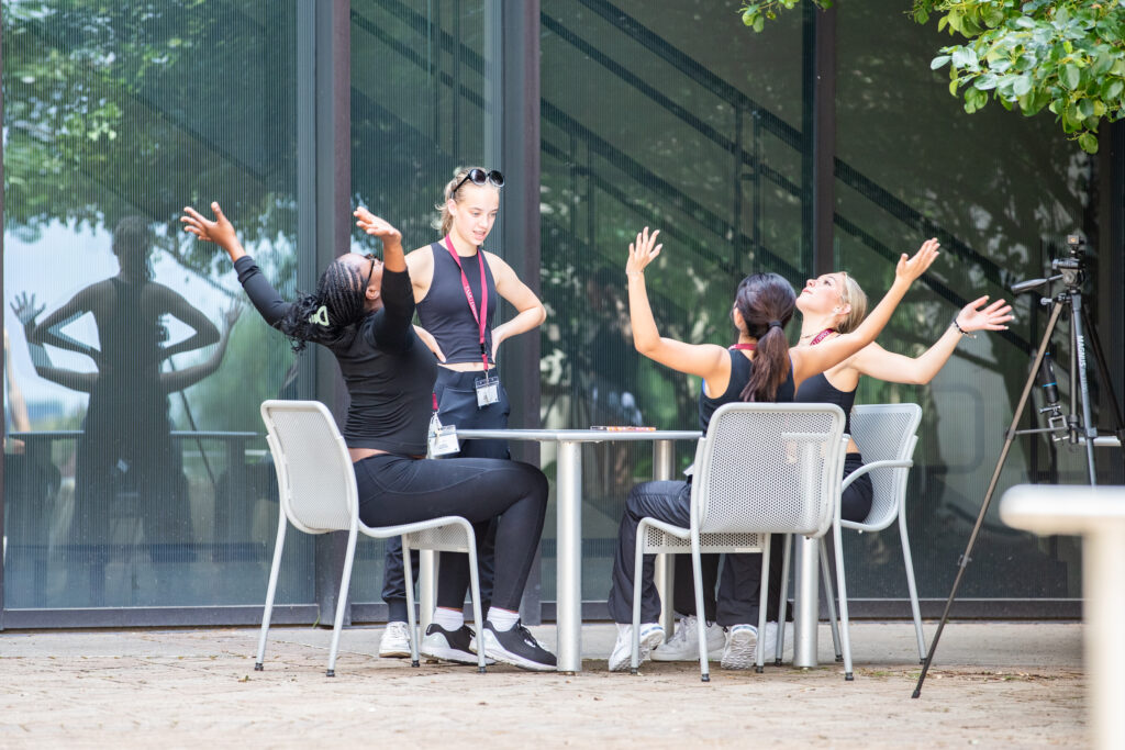 Three high school students seated at a table extend their arms up and out, while one standing student watches.