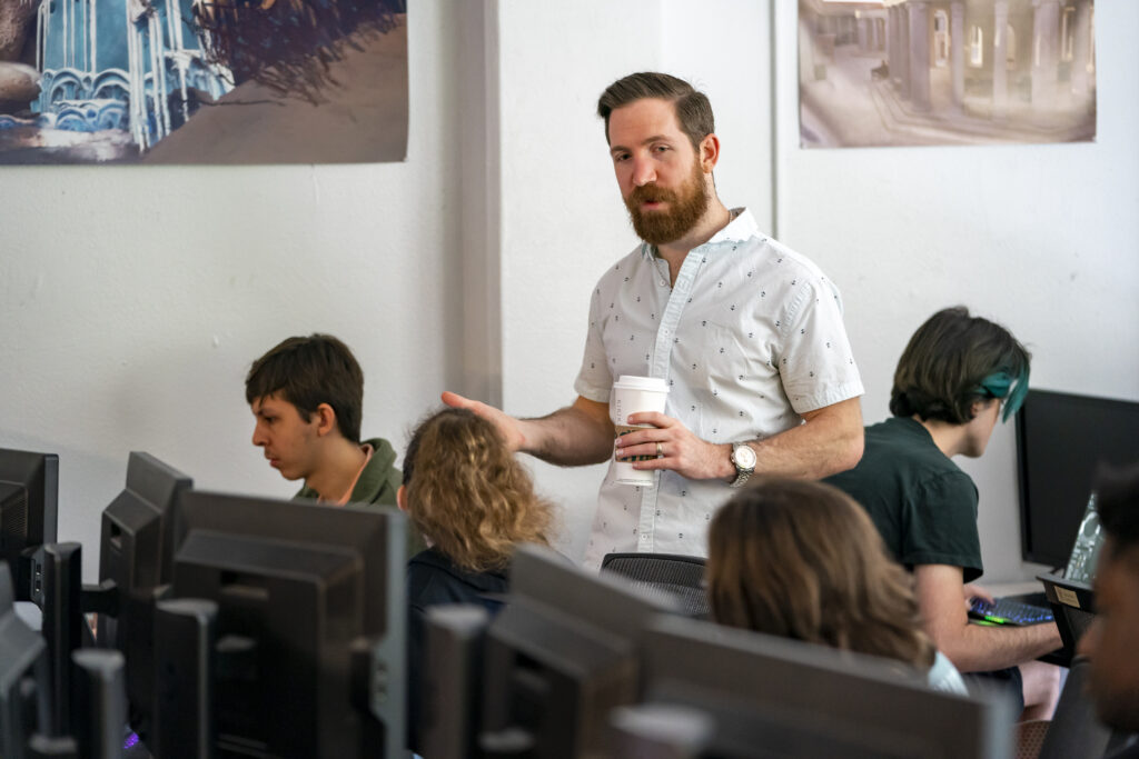 A game development professional speaks to college students who are seated at their desks and working on their games.