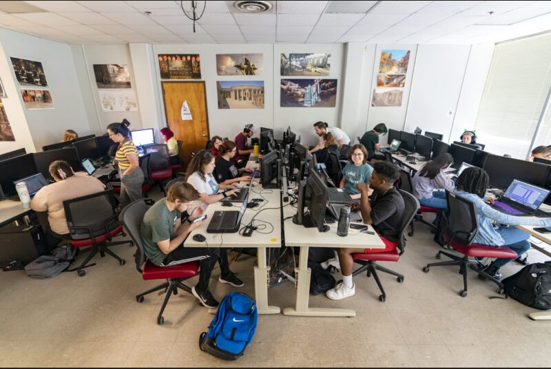 Students are seated at four long tables with computers as they work on their video game projects.