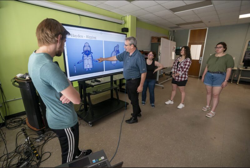 A college professor stands in front of a screen showing game characters in development. He gestures toward the screen while four students around him look on.