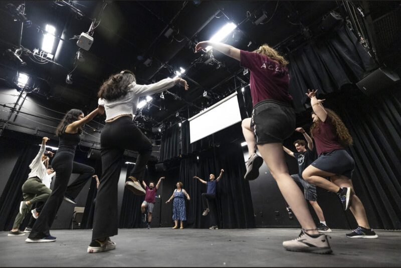 College students and staff members participate in an acting improv workshop, with people raising their arms and lifting one leg.