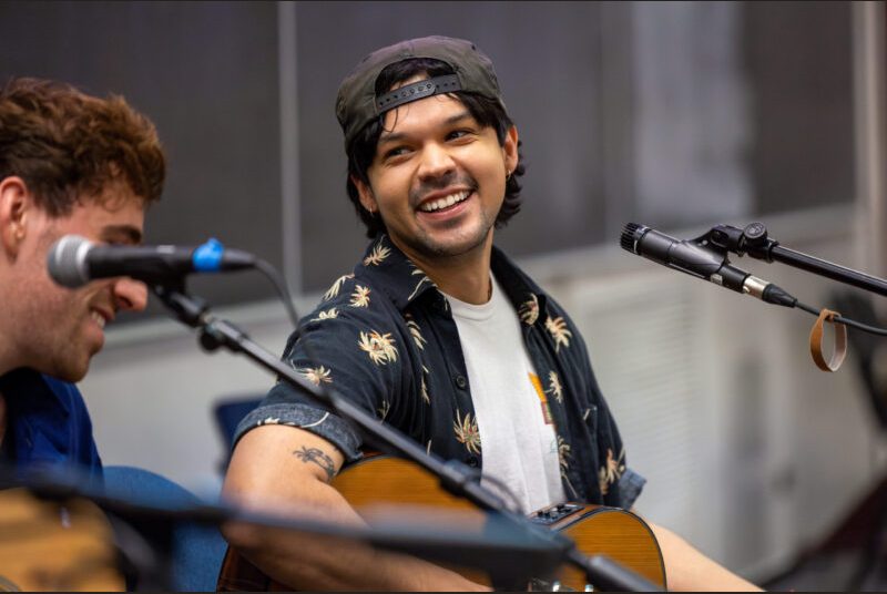 A member of a rock group smiles as he performs in a classroom in front of college students.