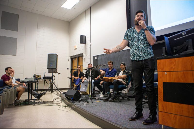 A man introduces a rock group in a classroom full of college students.
