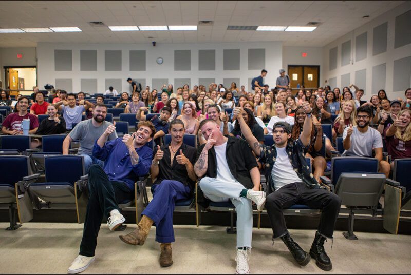 A rock group poses for a photo with college students in a classroom.