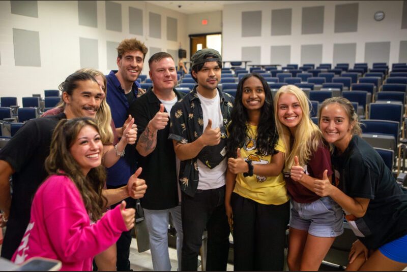 A rock group poses for a photo with college students in a classroom.