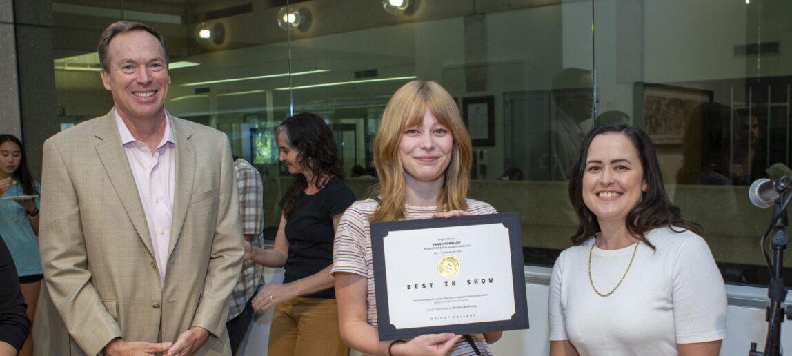A college student displays a certificate for earning Best in Show honors in an art exhibition. A professor stands on her left and a college dean stands on her right.