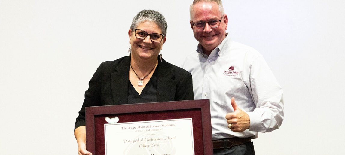 A college professor stands with a framed award while a university leader on her left gives the thumbs-up gesture.