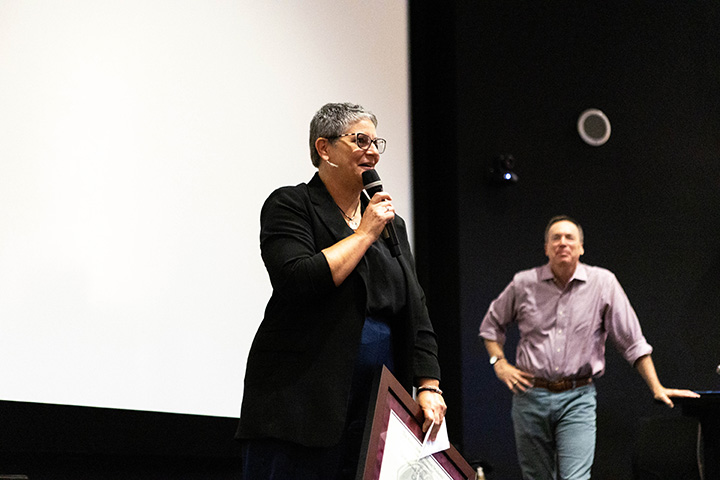 A college professor addresses an audience onstage while holding a framed award.