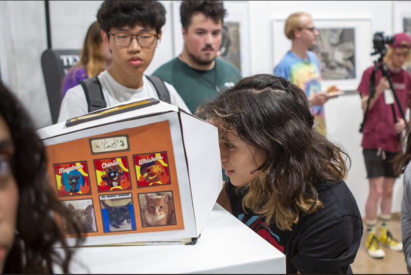 A college student looks through a small cutout in a box to view a three-dimensional diorama inside.