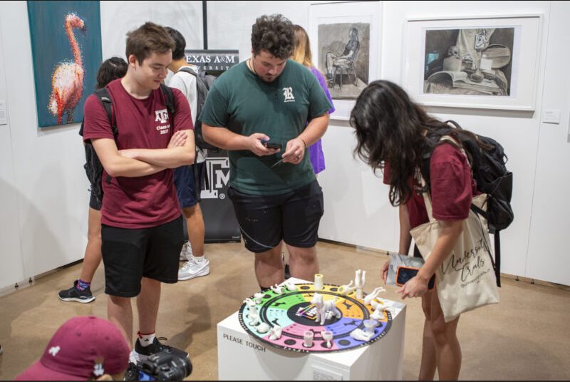 Three college students at an art gallery play with a board game about the characteristics of dogs.