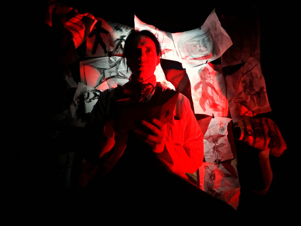 An image of a man standing in front of a wall of artwork, with red light surrounding him.
