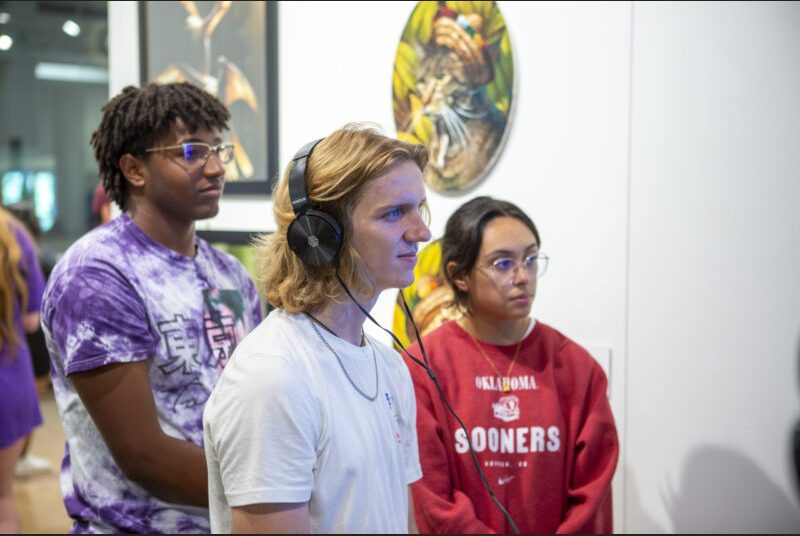 A college student wears headphones while watching a screen on the wall. Two students stand behind him.